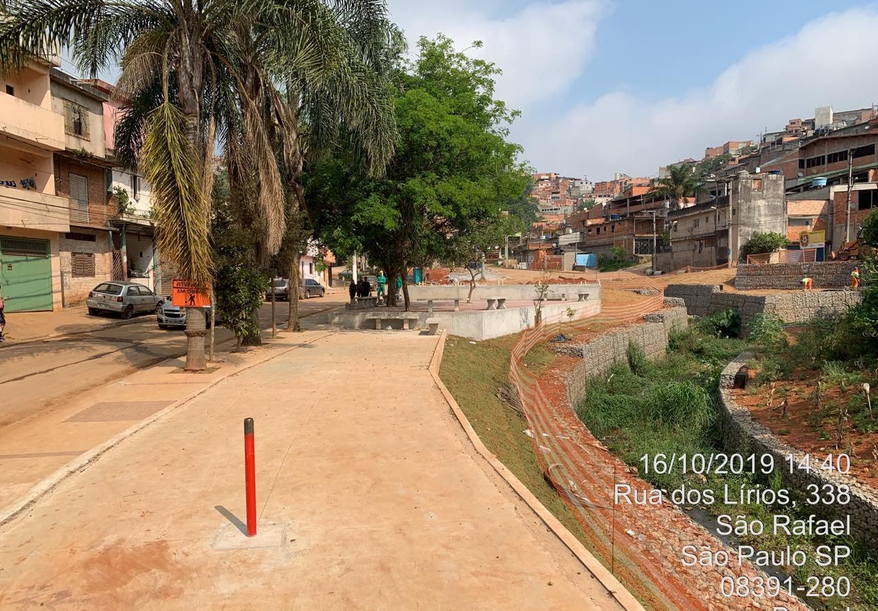 Passeio de concreto, margeando pequeno córrego, com muro de pedra nas margens. Nas laterais, casas humildes.